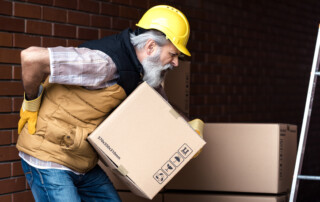 man with hard hat holding his lower back while lifting a box