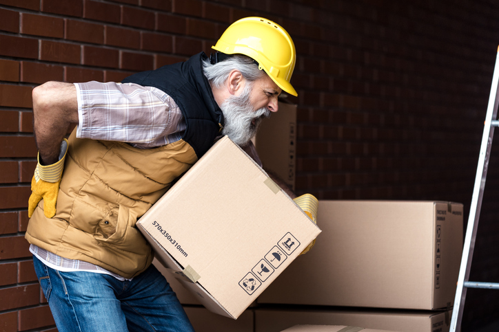 man with hard hat holding his lower back while lifting a box